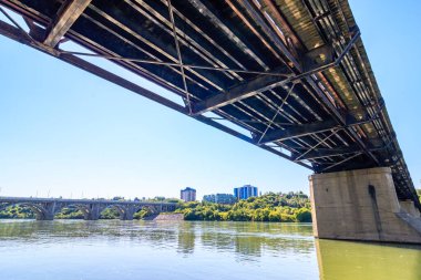 Bir köprü, arka planda bir şehir olan bir nehri kapsar. Köprü eski ve paslı, ama hala ayakta. Su sakin ve gökyüzü berrak. Huzurlu bir atmosfer yaratıyor.