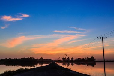 Arka planda elektrik hattı olan bir gölün üzerinde güzel bir günbatımı. Gökyüzü bulutlarla dolu ve su sakin.