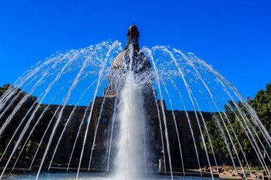 Binanın önünde içinden su fışkıran bir çeşme. Su beyaz ve gökyüzü mavi.
