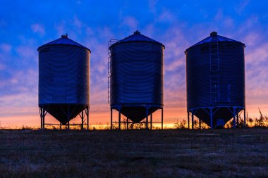 Gün batımında üç büyük siyah tank bir tarlada oturur. Gökyüzü turuncu ve mavi renklerin karışımıdır.