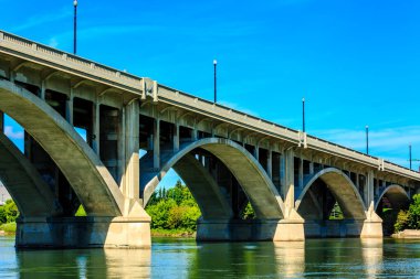 Üstü açık mavi gökyüzü olan bir nehir boyunca uzanan bir köprü. Köprü betondan yapılmıştır ve modern bir tasarıma sahiptir.