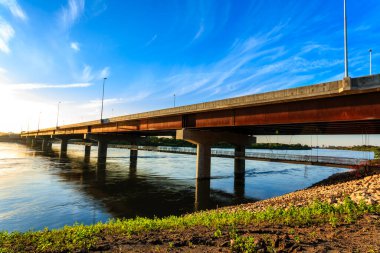 Üstü açık mavi gökyüzü olan bir nehir boyunca uzanan bir köprü. Köprü çimenler ve kayalarla çevrili ve su sakin.