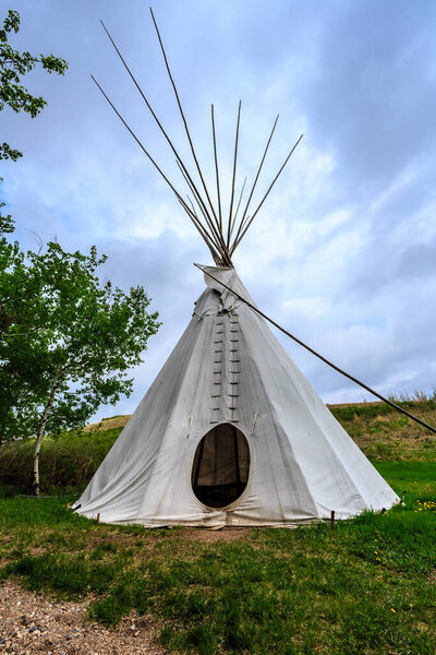 A teepee is standing in a grassy field. The teepee is white and has a pointed roof. The sky is cloudy and the grass is green