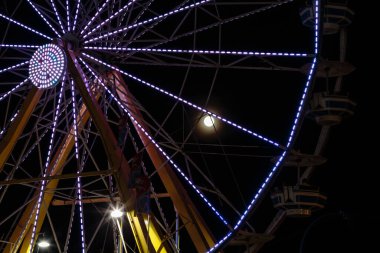 Arka planda dolunay olan büyük, aydınlık bir dönme dolap. Ay ışıl ışıl parlıyor, direksiyonun üzerine sıcak bir parıltı saçıyor. Sahne huzurlu ve huzurlu.