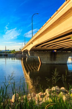 Bir köprü, sudaki gökyüzünün bir yansımasıyla bir nehri kaplar. Köprü betondan yapılmıştır ve çimen ve kayalarla çevrilidir.