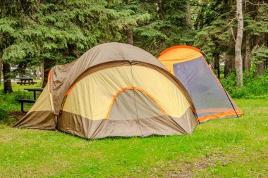 Çimenli bir alanda büyük bir çadır kurulur. Çadır ağaçlarla çevrili ve yakında bir piknik masası var. Sahne huzurlu ve davetkâr. Kamp gezisi için mükemmel.