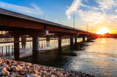 Köprü, arka planda günbatımı olan bir nehri kapsar. Köprü betondan yapılmıştır ve kayalarla çevrilidir.