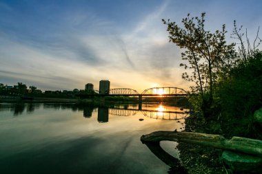 Bir köprü, arka planda bir şehir olan bir nehri kapsar. Güneş batıyor, sahne üzerine sıcak bir parıltı saçıyor.