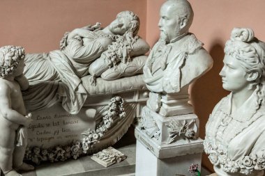 Bir erkek ve bir kadın heykeli bir yatakta oturuyor. Adam şapka takıyor ve kadın da elbise giyiyor. Heykel çiçeklerle çevrili.