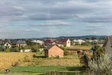 Küçük tuğlalı bir ev ve ahır olan bir kırsal bölge. Gökyüzü bulutlu ve evler yayılmış.