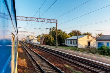Bir tren mavi beyaz bir tren vagonuyla raylarda ilerliyor. Tren birkaç evi ve kilisesi olan küçük bir kasabadan geçiyor. Gökyüzü açık ve güneş parlıyor.