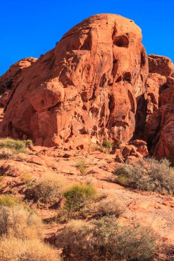 Arka planda mavi gökyüzü olan kayalık bir dağ. Dağ kırmızı toprakla kaplıdır ve üzerinde birkaç çalı yetişir.