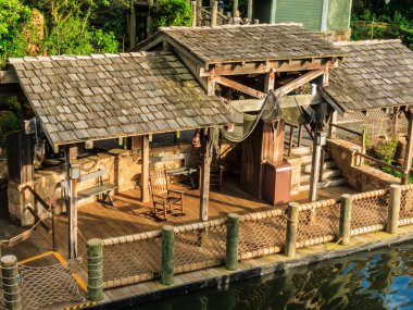 Ahşap bir yapı, eğimli bir çatı ve ahşap bir güverte. Güvertede bir sandalye ve bir bank var. Bina bir çitle çevrili ve bir su kütlesinin yakınında.