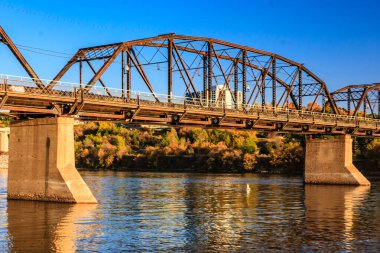 Bir köprü, arka planda bir şehir olan bir nehri kapsar. Köprü eski ve paslı, ama hala ayakta. Su sakin ve uzaktaki köprüyü ve şehri yansıtıyor.