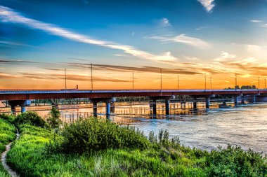 Bir köprü arka planda güzel bir günbatımı olan bir nehri kapsar. Köprü kırmızı ve beyaz ve su sakin. Gökyüzü bulutlarla dolu. Huzurlu ve huzurlu bir atmosfer yaratıyor.