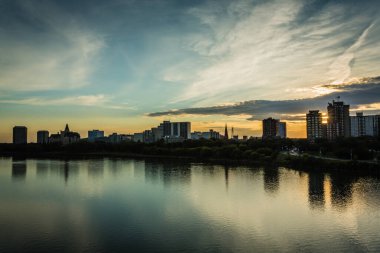 Gün batımında sakin bir göle şehrin ufuk çizgisi yansıtılır. Gökyüzü bulutlarla dolu. Huzurlu ve huzurlu bir atmosfer yaratıyor.