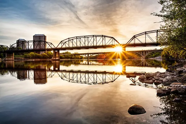 Köprü, arka planda günbatımı olan bir nehri kapsar. Köprü eski ve uzun bir geçmişi var.