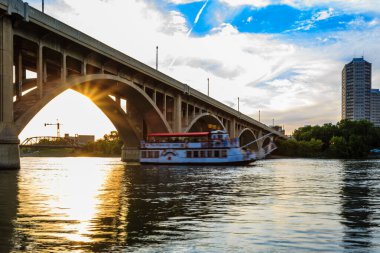 Bir tekne nehrin üzerindeki köprüden geçiyor. Güneş batıyor, suyun üzerine sıcak bir parıltı saçıyor.
