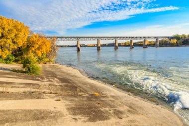 Köprü, diğer tarafında kumlu bir plaj olan bir nehri kapsar. Gökyüzü mavi ve su sakin.