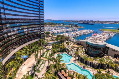 Arka planda büyük bir su kütlesi ve havuzu olan bir otel. Otel palmiye ağaçları ile çevrilidir ve büyük bir çeşmesi vardır.