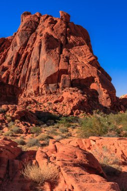 Üzerinde açık mavi gökyüzü olan büyük bir kırmızı kaya oluşumu çölde oturmaktadır. Sahne huzurlu ve sakin, kaya oluşumu çevredeki manzaranın üzerinde yükseliyor.