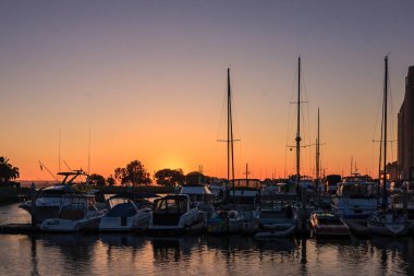 Güneşin arka planda battığı, bir grup botun kenetlendiği sakin bir marina sahnesi. Teknelerin boyutları ve renkleri farklı. Su sakin ve durgun.