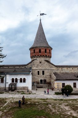 Bir grup insan çan kulesiyle büyük bir binanın etrafında yürüyorlar. Bina eski ve bir sürü tarihi var. Gökyüzü bulutlu ve atmosfer biraz kasvetli.
