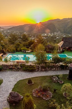 Güzel bir havuz manzarası ve arka planda günbatımı olan bir dağ. Havuz ağaçlar ve taş bir duvarla çevrili.