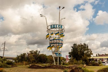 Üzerinde ODEK yazan uzun, çarpık, mavi ve sarı bir tabela. Tabela ağaçlarla çevrili ve bir tarlada bulunuyor.