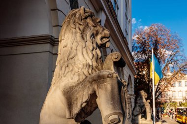 Bir binanın önünde bir aslan heykeli duruyor. Heykel taştan yapılmıştır ve büyük bir yelesi vardır. Bina yüksek ve bir sürü penceresi var. Gökyüzü mavi ve birkaç bulut var.