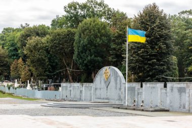 Arka planda büyük bir bayrak dalgalanan bir mezarlık. Bayrak sarı ve mavi. Mezarlık bir duvarla çevrili.