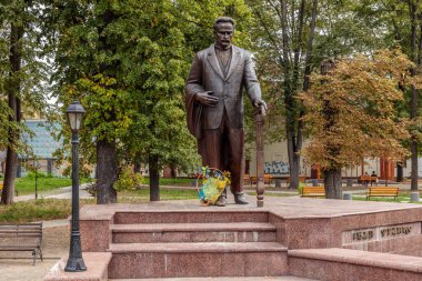 Elinde bir buket çiçek olan bastonlu bir adam heykeli. Heykel, ağaçlı ve banklı bir parkta bulunuyor.