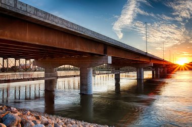Köprü, arka planda günbatımı olan bir nehri kapsar. Köprü betondan yapılmıştır ve büyük bir kemere sahiptir. Su sakin ve gökyüzü bulutlarla dolu.