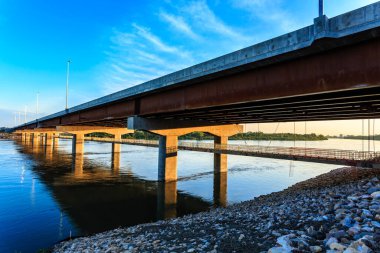 Bir köprü aşağıdaki su manzaralı bir nehri kapsar. Köprü betondan yapılmıştır ve üzerinde birkaç masa ve sandalye vardır. Su sakin ve gökyüzü mavi.