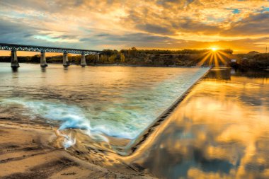 Köprü, arka planda günbatımı olan bir nehri kapsar. Su sakin ve gökyüzü bulutlarla dolu.
