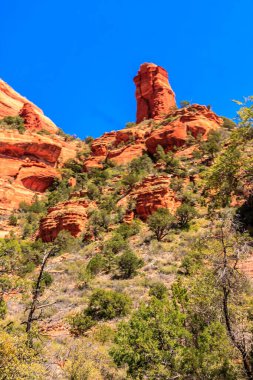 Tepesinde büyük bir kaya olan ve yanında ağaçlar büyüyen bir dağ. Gökyüzü mavi ve güneş parlıyor.