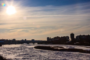 Arka planda içinden bir nehir akan bir şehir silueti görülebilir. Gökyüzü bulutlu ve güneş parlıyor.
