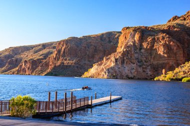Bir adam rıhtımın yakınındaki bir gölde jet ski sürüyor. Gökyüzü mavi ve su sakin.