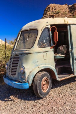 Eski mavi beyaz bir kamyon çakıllı bir yolda park halinde. Kamyon paslanmış ve soluk mavi bir boyası var. Kamyon kayalık bir tepenin önünde park halinde.