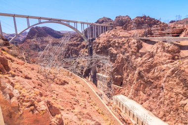 Bir köprü, arka planında kayalık bir dağ sırası olan bir kanyonu kapsar. Köprü, büyük kemerli bir asma köprüdür.