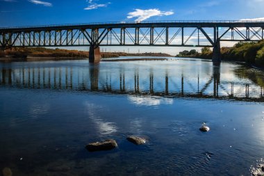 Bir köprü, sudaki gökyüzünün bir yansımasıyla bir nehri kaplar. Köprü metalden yapılmıştır ve ağaçlarla çevrilidir.