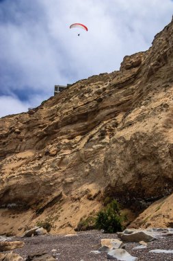 Bir insan kayalık bir uçurtmanın üzerinde gökyüzünde uçurtma uçuruyor. Sahne sakin ve huzurlu, uçurtma kayalık arazide süzülüyor.