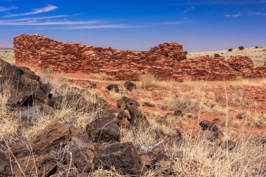 Uzakta büyük bir taş bina ile kayalık bir çöl manzarası. Gökyüzü açık ve mavidir, ve güneş parlıyor. Sahnenin yalnızlık ve yalnızlık hissi var.