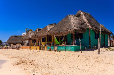 Sazlı çatıları olan renkli evlerle dolu bir sahil. Evler parlak renklerle boyanmış ve tropikal bir hissi var. Kumsal boş, görünürde kimse yok.