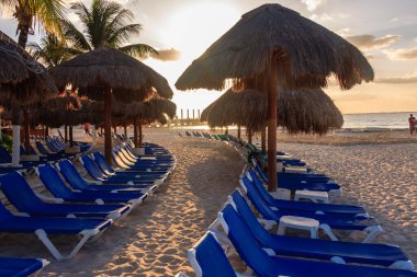 Mavi şezlonglar ve şemsiyelerle dolu bir kumsal. Kumsal boş ve güneş batıyor.