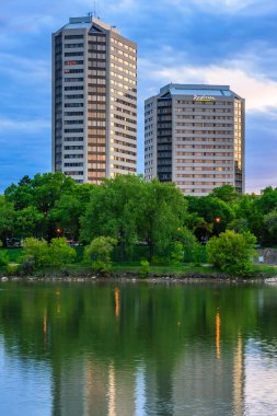 İkisinin arasında büyük bir su kütlesi olan iki yüksek bina. Su sakin ve binaları yansıtıyor.