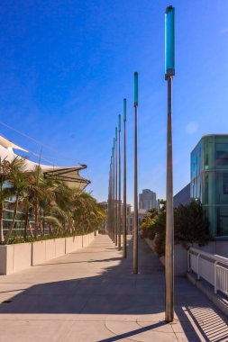 Üzerinde mavi bir gökyüzü ve arkasında palmiye ağaçları olan uzun bir yürüyüş yolu. Geçit uzun direklerle kaplı ve sağ tarafında beyaz bir çit var.