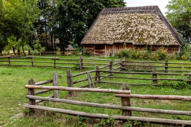 Ahşap bir çit sazdan çatılı küçük bir evi kuşatır. Ev yemyeşil bir tarla ile çevrili.