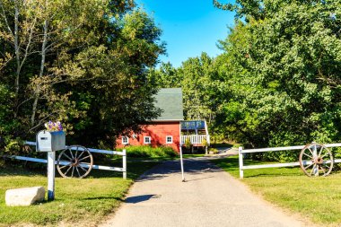 Beyaz posta kutusu ve ahşap çiti olan kırmızı bir ahır. Ahır ağaçlarla çevrili ve yol asfalt kaplı.