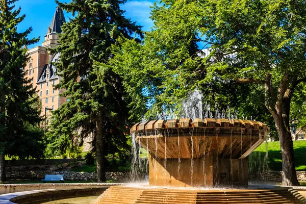 Parkta içinden su fışkıran büyük bir çeşme. Çeşme ağaçlarla çevrili ve arka planda bir bina var. Sahne huzurlu ve sakin, suyun sesiyle.